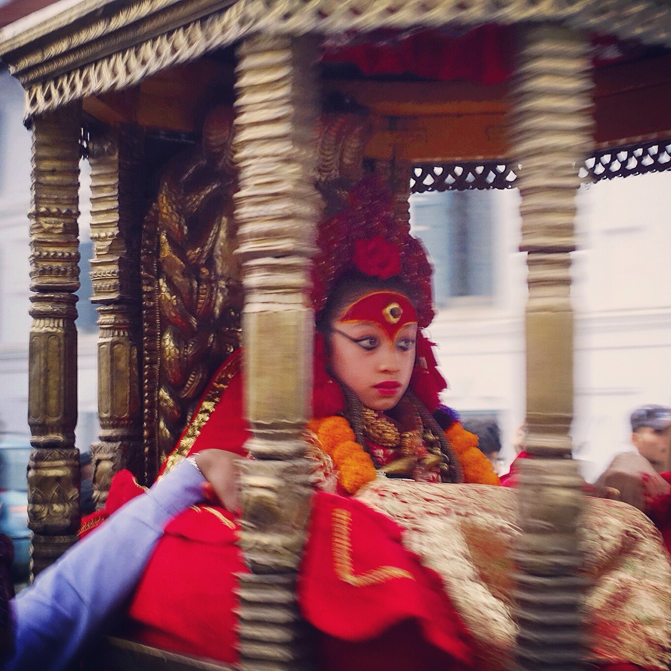 A young girl wearing red robes and headdress and heavily made up sitting in a palaquin