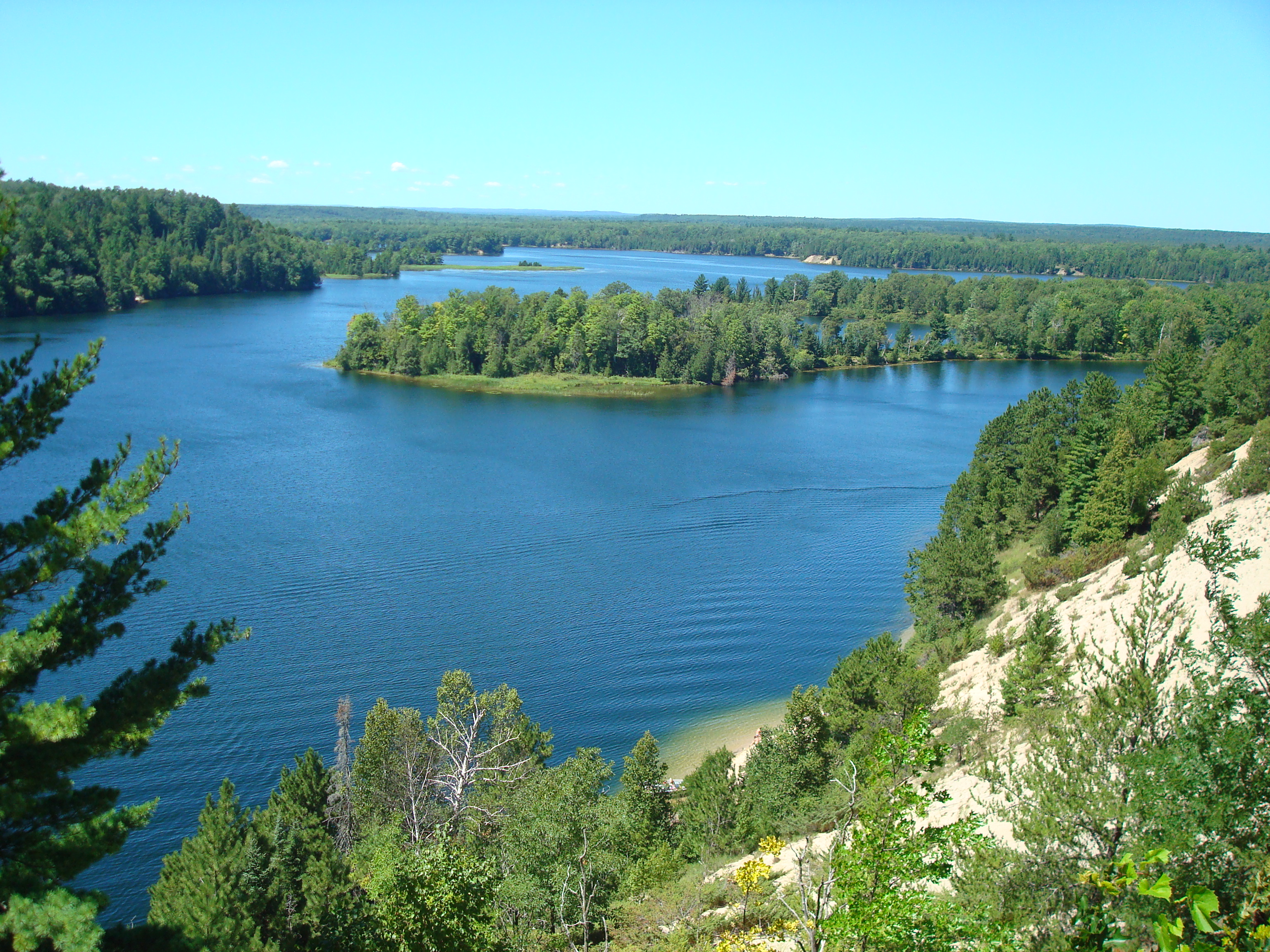 View of a blue river under a clear blue sky with a green island in the middle and a steep sand dune in the foreground