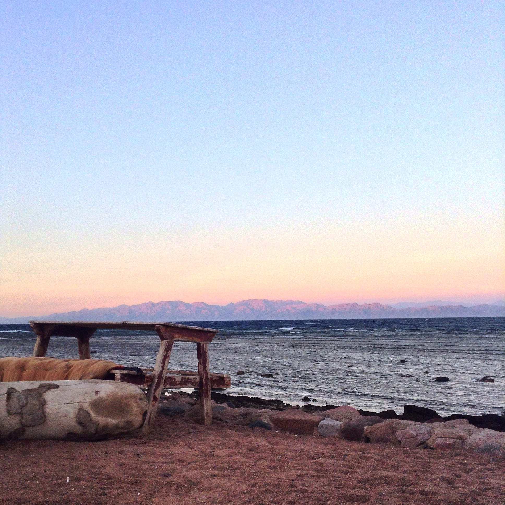 A picnic table on a beach with the pastel colors of a sunset in the background with mountains on the other side of the Red Sea