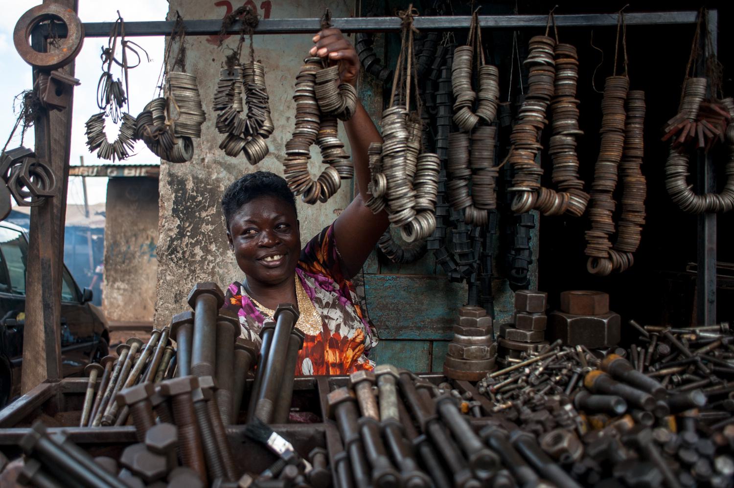 a woman standing behind a table covered in nuts and bolts, smiling and reach up to a loop of string covered with metal washers