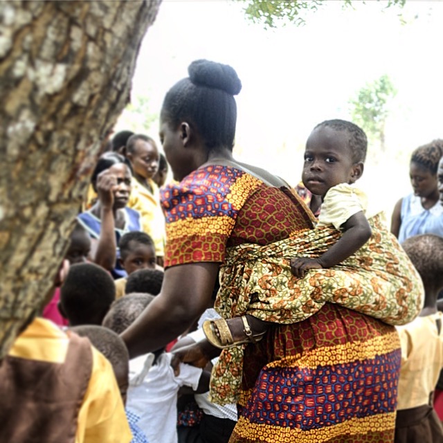 a woman bending slightly at the waist with a baby, who is looking at the camera, tied on her back using a length of fabric