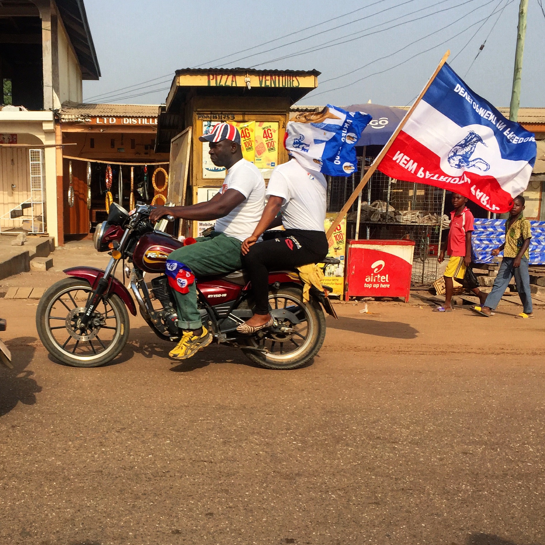 A motorcycle with two riders drives by with the Ghanaian National Patriotic Party flag waving from behind