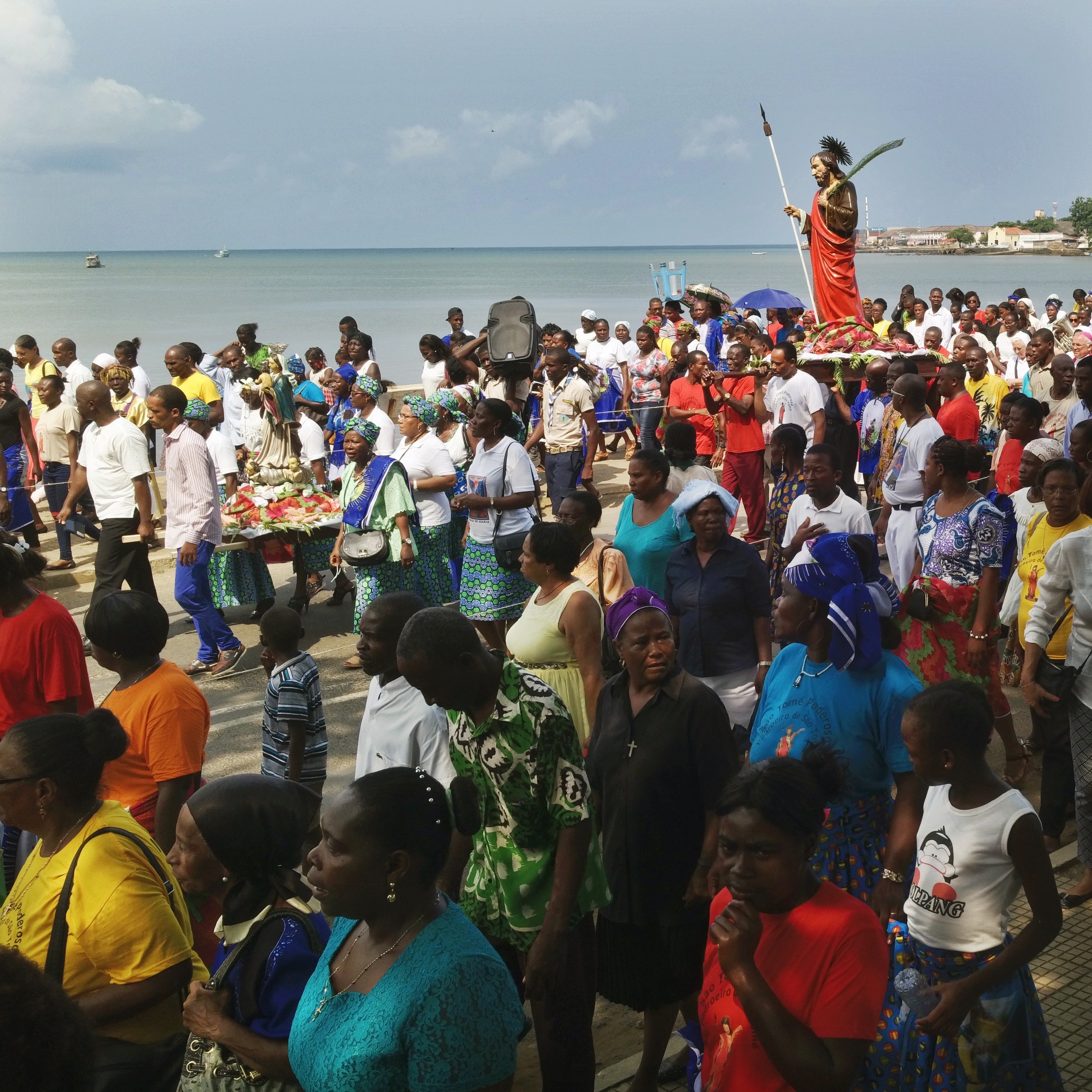 a crowd of people around a statue of st thomas parading on a street in front of the ocean