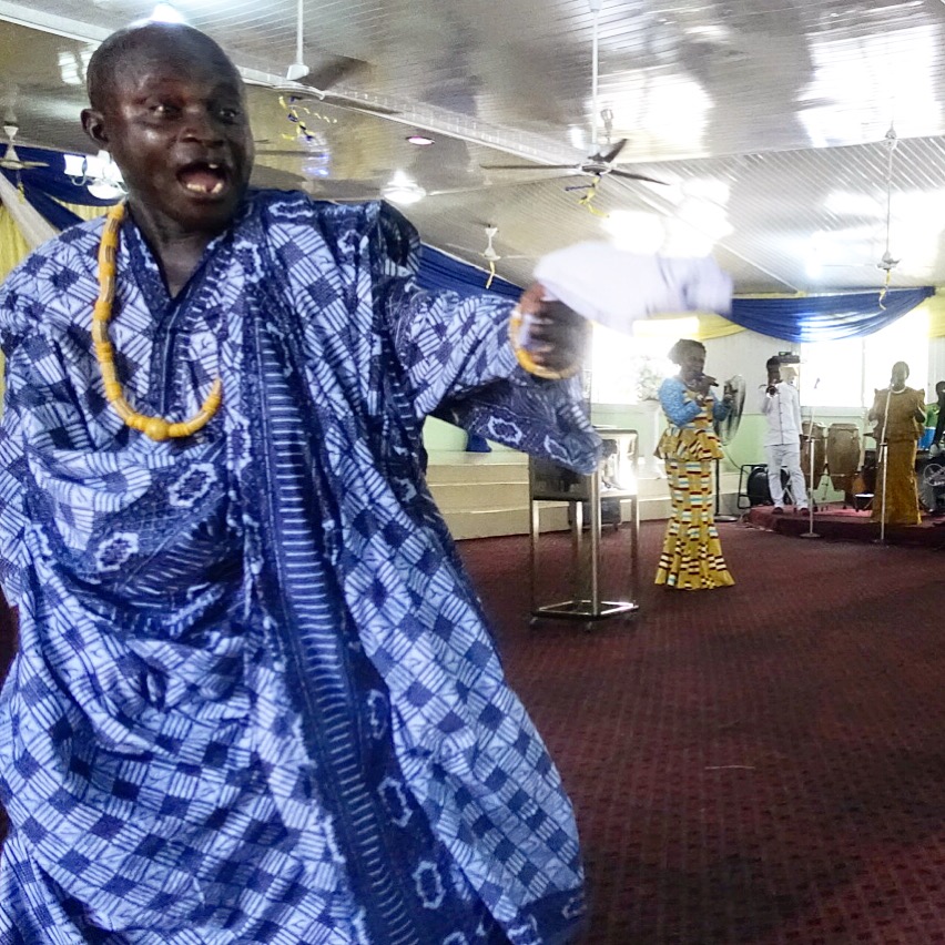 A man wearing a light blue and dark blue patterned cloth dances with a large smile.