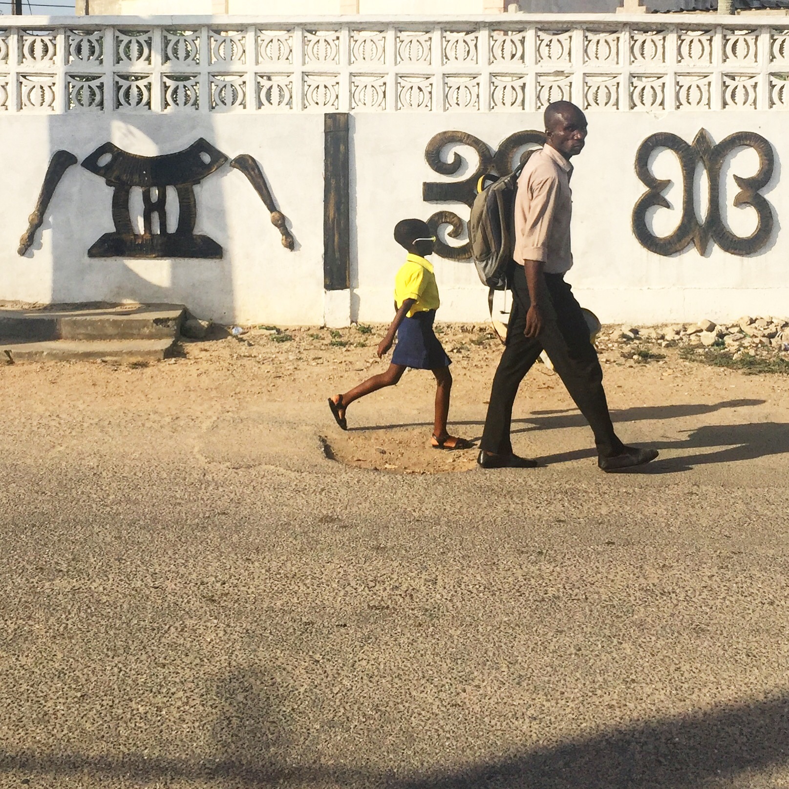 A man and young girl walking in front of a white wall with black adinkra symbols on it.