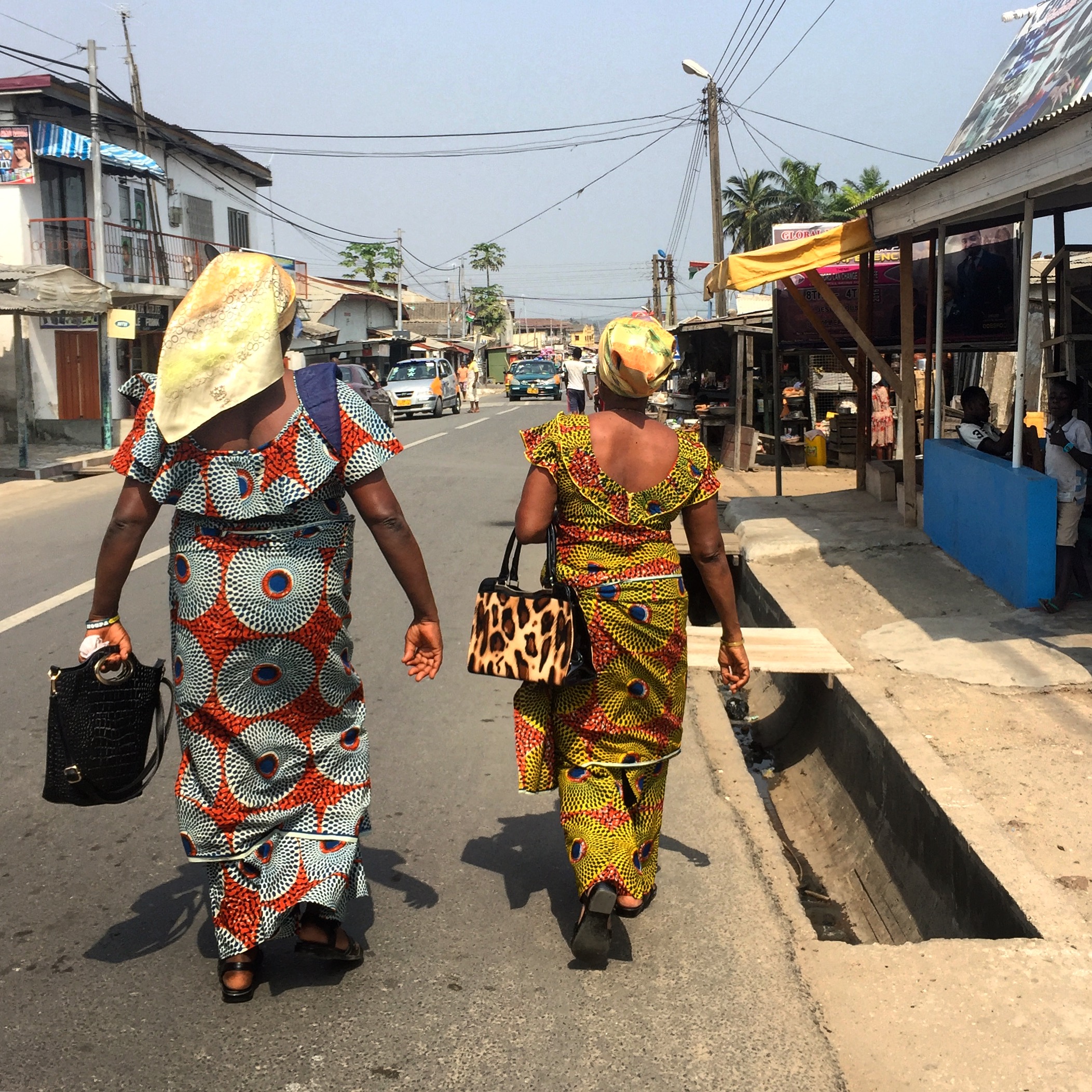 Two women walking away from the viewer on a street wearing the traditional Ghanaian outfit of a kaba and slit in African print.