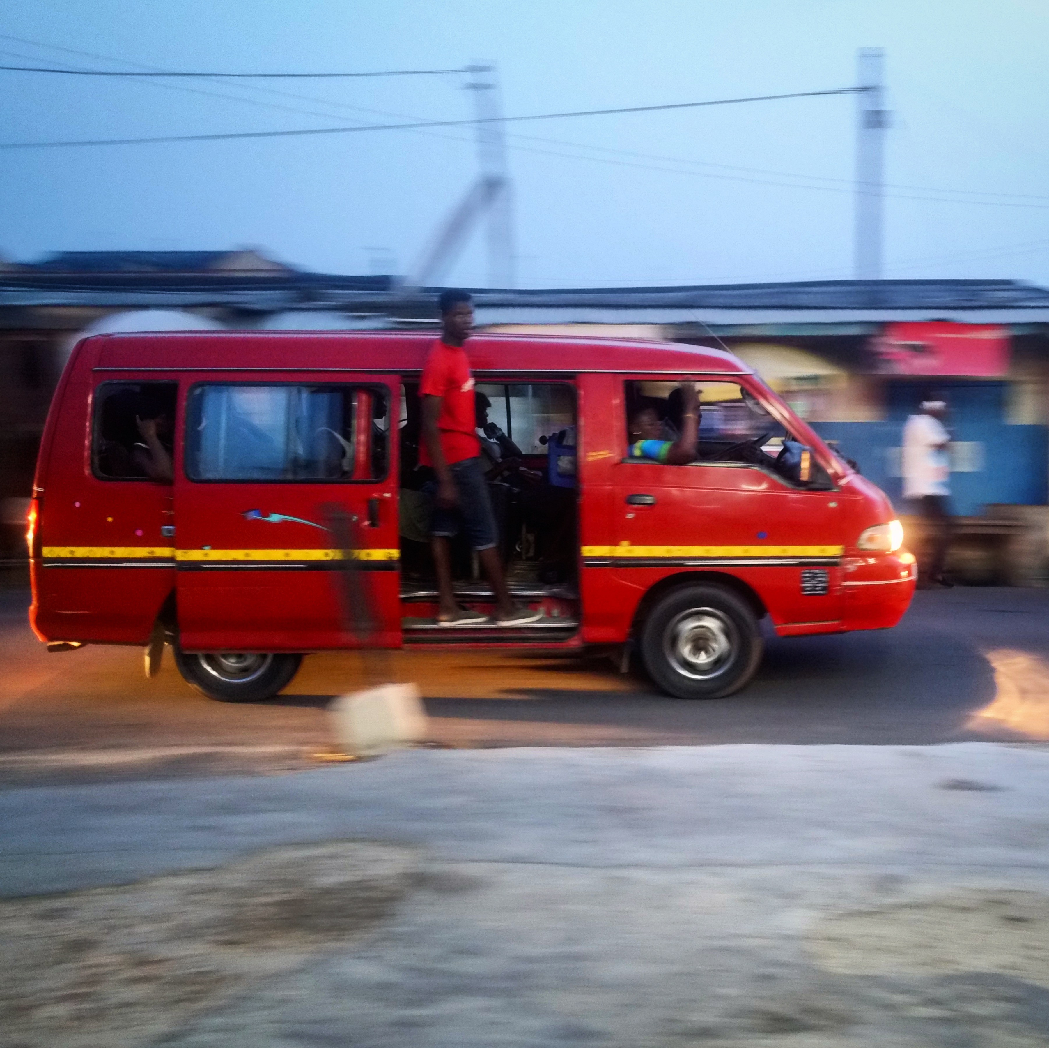 A red van, with the side door open and a young man standing and hanging off the vehicle, is moving from left to right.