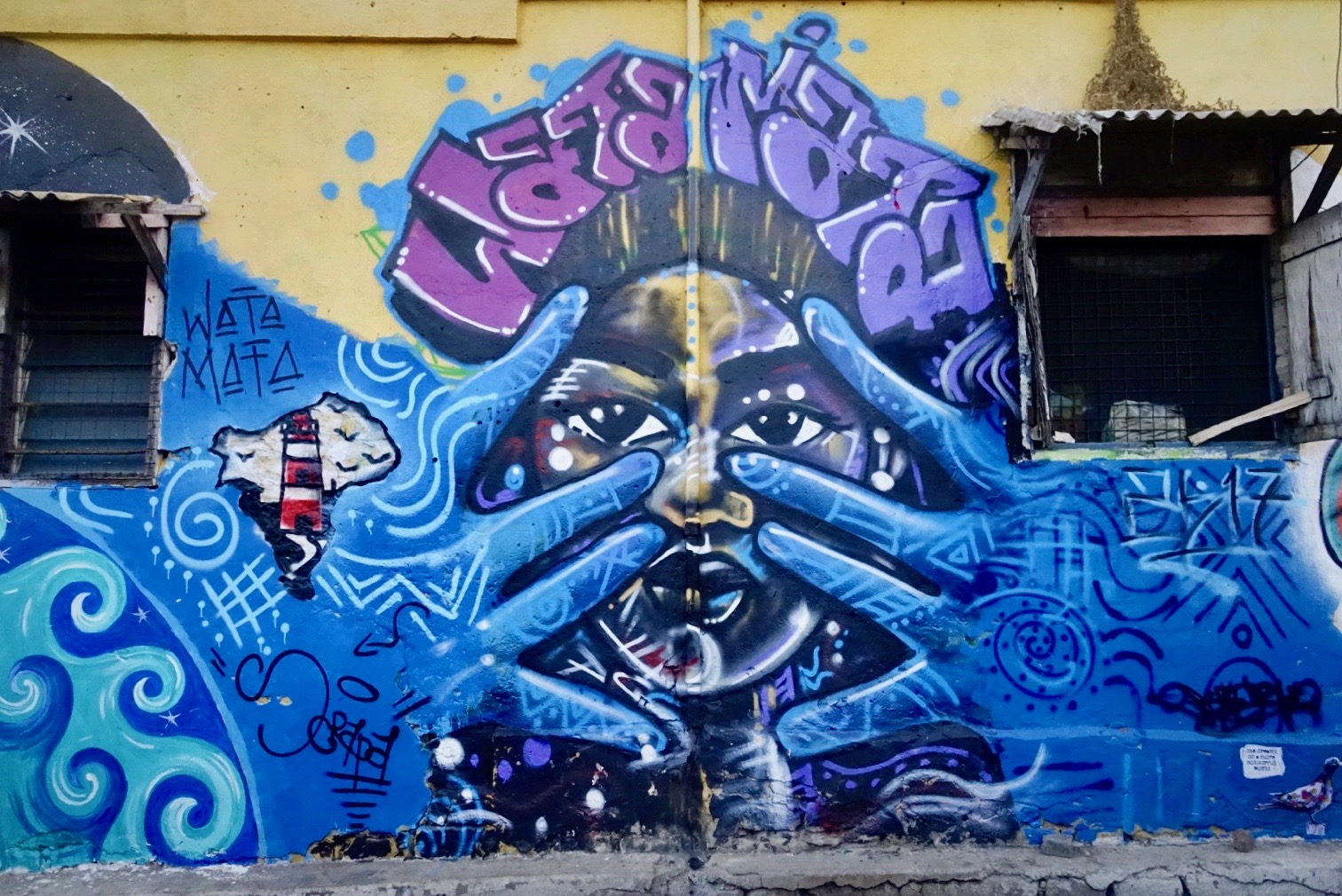 A mural of a woman with blue hands held in front of her face under the words "Wata Mata"