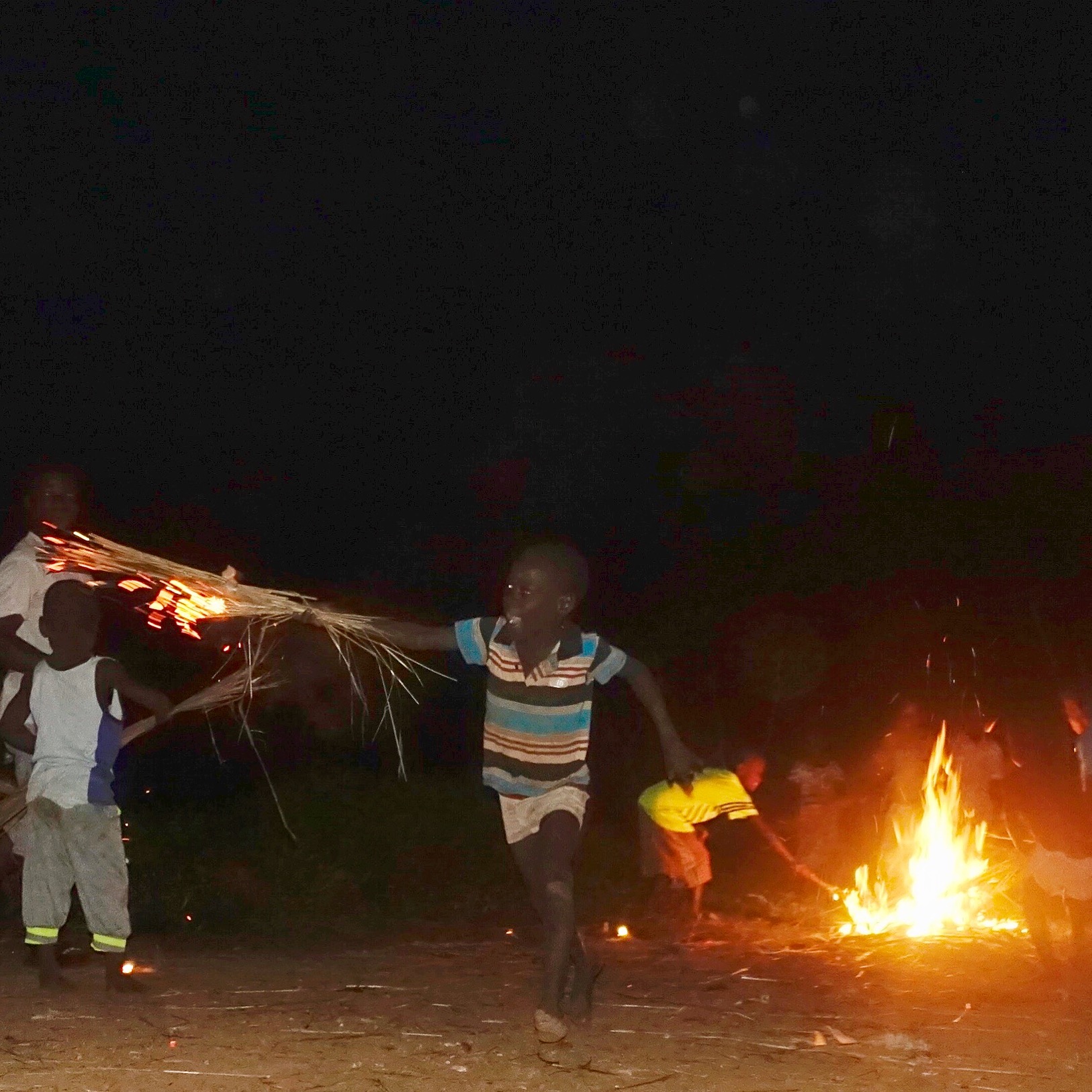 a small boy, holding a bundle of grass that is lit on fire runs towards the camera and a fire burns in the background under a black sky